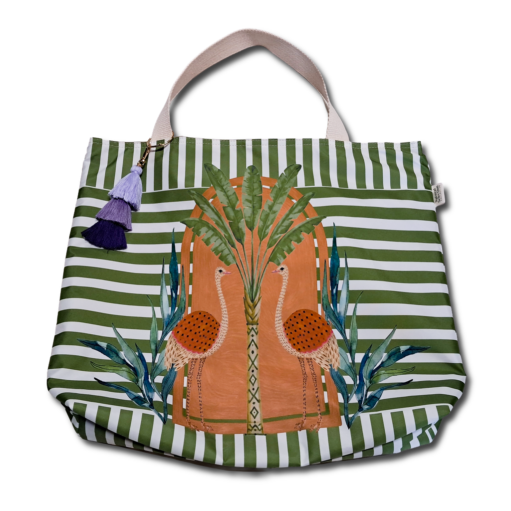 A canvas tote bag with green and white horizontal stripes, featuring a central illustration of two flamingos in front of a palm tree. The bag has light beige shoulder straps and a decorative purple tassel charm on the left side, shown against a plain white background.