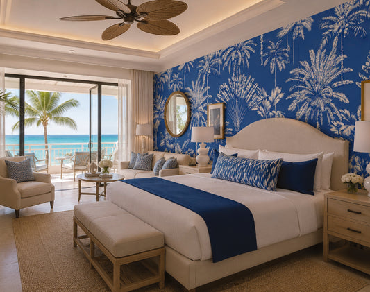 A luxury bedroom featuring a large bed with white linens and a royal blue runner, set against a bold blue and white palm tree patterned wall. The room includes a matching blue accent pillow, a beige upholstered bench at the foot of the bed, a matching armchair, and a large window overlooking a tropical beach with palm trees.