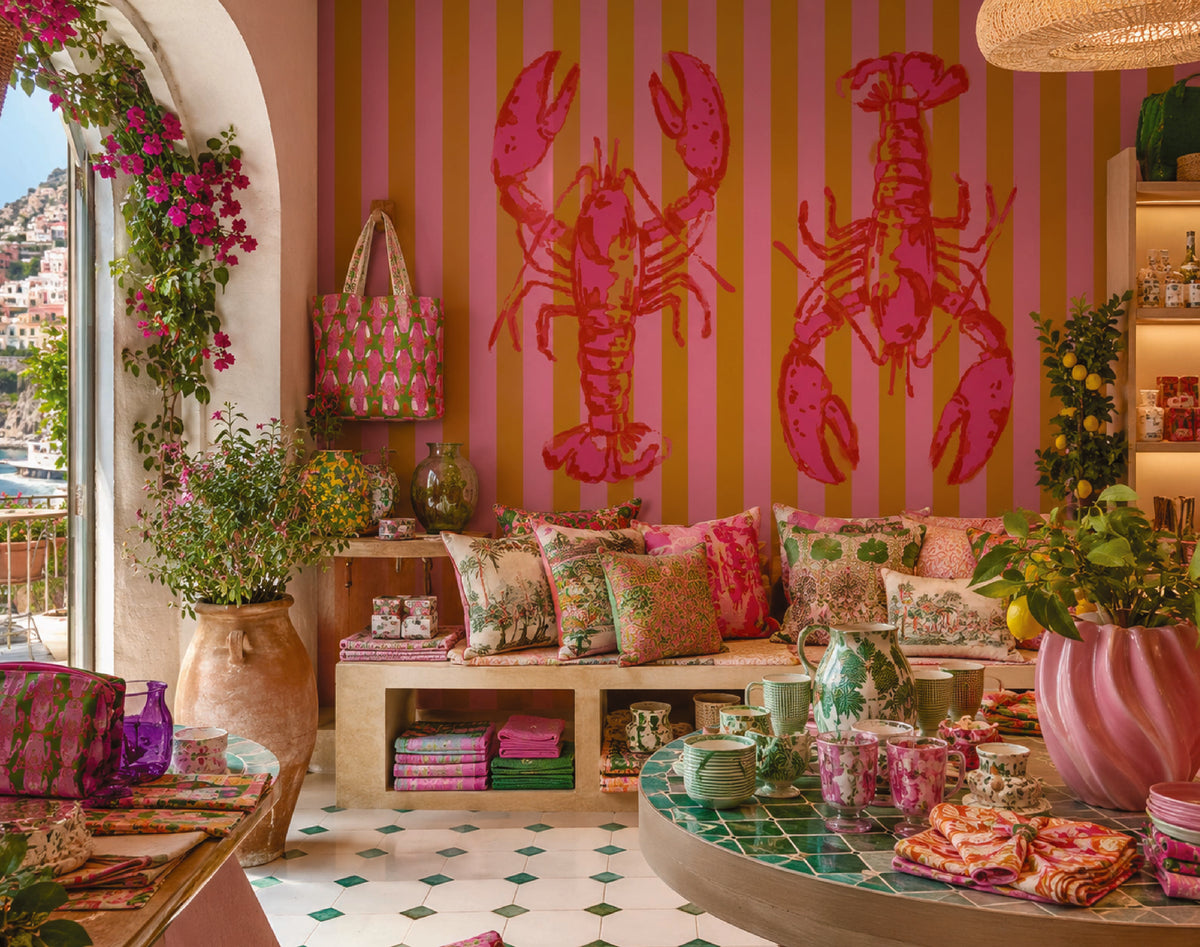 A vibrant, highly styled interior room featuring a bold pink and orange striped wall with large lobster silhouettes, a wooden bench with patterned pillows, and a green-tiled coffee table in the foreground.