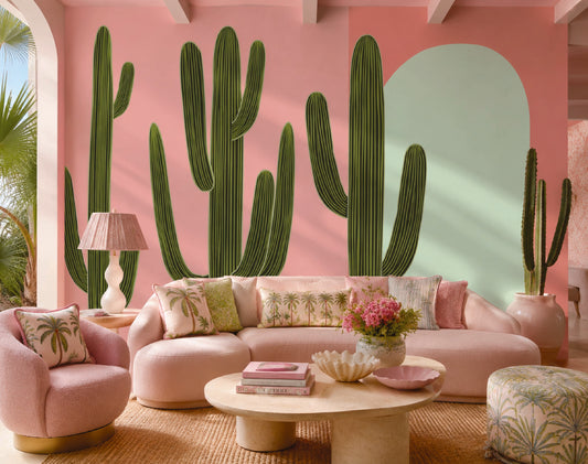 A styled living room featuring a large pink wall mural with green cactus silhouettes and a mint green arch. A curved pink sofa with patterned pillows, a matching pink armchair, a round wooden coffee table, and a patterned ottoman are arranged in front of the mural.