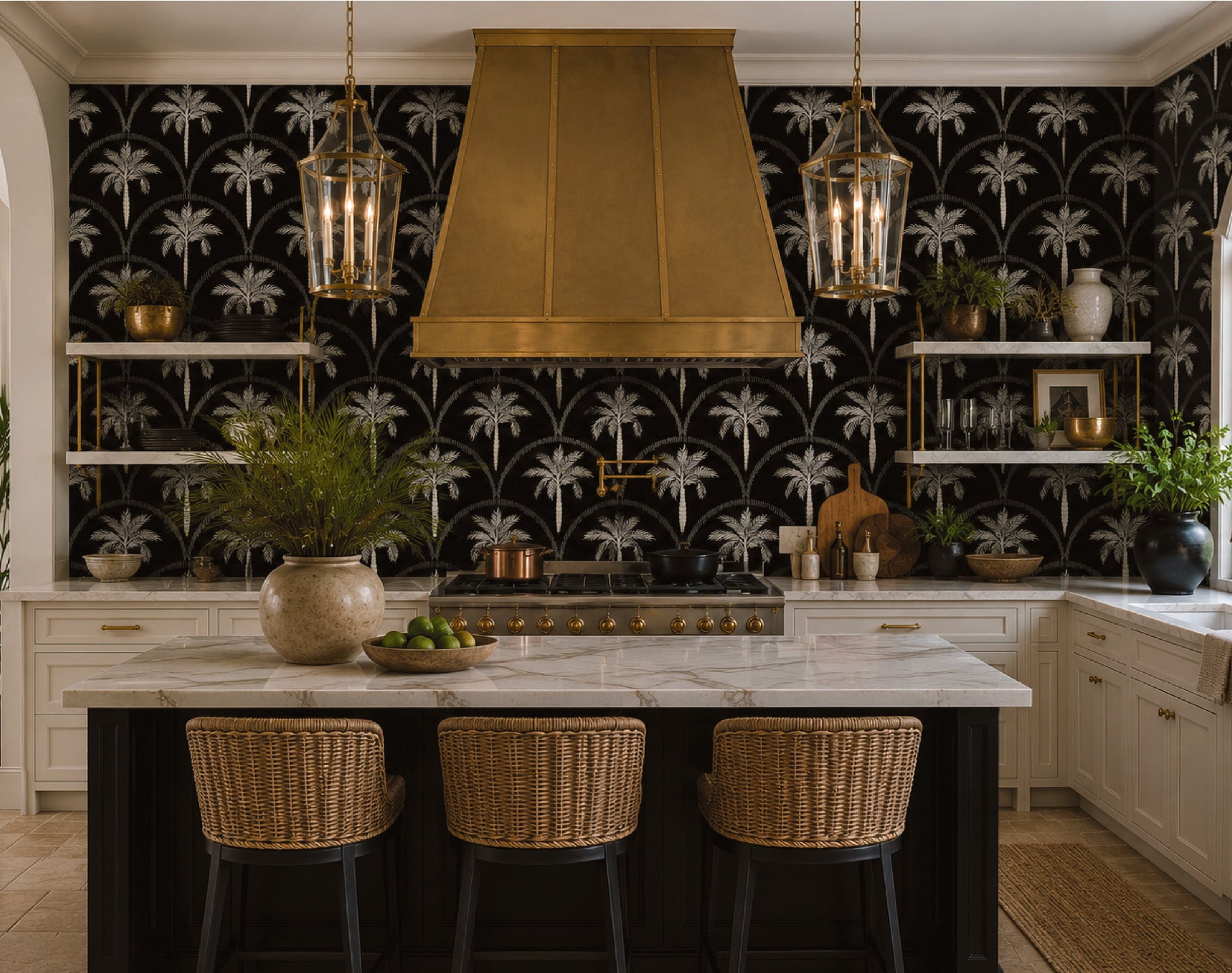 A luxury kitchen interior featuring a bold black and white palm tree patterned wallpaper, a large brass range hood, a marble-topped island with three woven bar stools, and hanging lantern pendant lights.