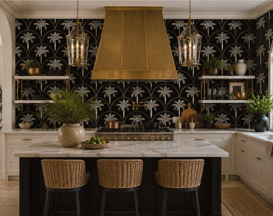 A luxury kitchen interior featuring a bold black and white palm tree patterned wallpaper, a large brass range hood, a marble-topped island with three woven bar stools, and hanging lantern pendant lights.