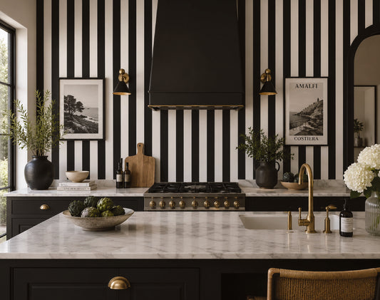 A modern kitchen interior with bold black-and-white vertical striped wallpaper, a black range hood, dark cabinetry, a white marble countertop, and brass fixtures. Framed wall art, plants, and decorative objects are arranged on the counters.