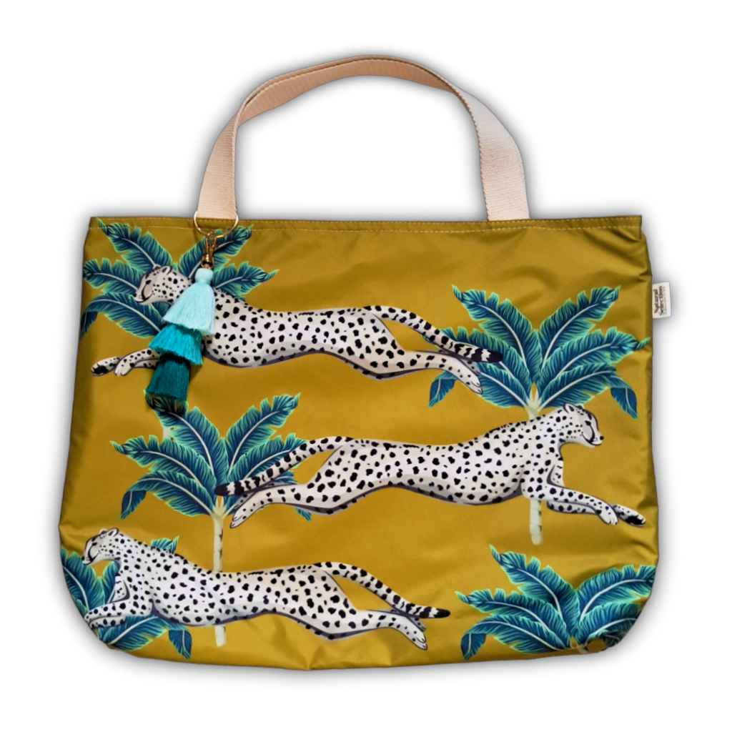 A mustard yellow tote bag featuring a pattern of leaping spotted cheetahs and blue-green palm trees, with beige shoulder straps and a hanging tassel charm.