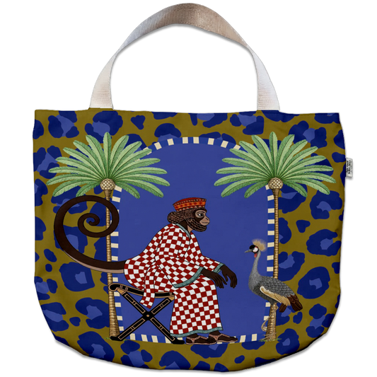 A tote bag featuring a vibrant, illustrated design of a seated monkey in a red-and-white checkered robe between two palm trees, set against a blue and gold leopard print background. The bag has a cream-colored fabric handle and a small side tag.
