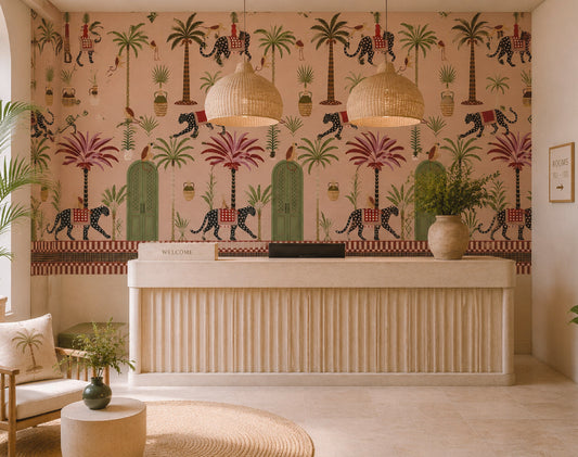 A styled interior reception area featuring a large wall mural with a whimsical safari print of palm trees, leopards, and green arches on a pink background. A cream-colored fluted reception desk sits in front of the wall, flanked by two woven pendant lights, a chair with a palm-print pillow, and potted plants.