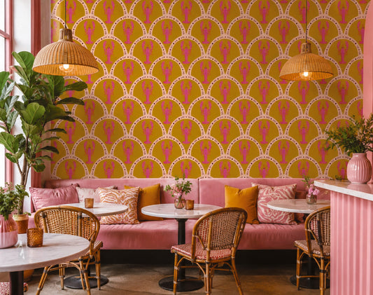 A vibrant interior scene featuring a bold wallpaper pattern with pink silhouettes on a mustard yellow background, styled with a pink velvet banquette, wicker chairs, and pendant lights.