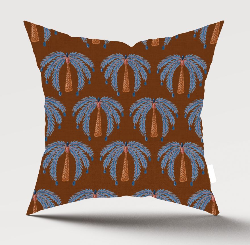 A square throw pillow featuring a repeating palm tree pattern on a warm brown background. The palm trees are light blue with orange trunks, and small blue hanging ornaments are visible.