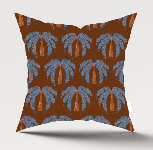 A square throw pillow featuring a repeating palm tree pattern on a warm brown background. The palm trees are light blue with orange trunks, and small blue hanging ornaments are visible.