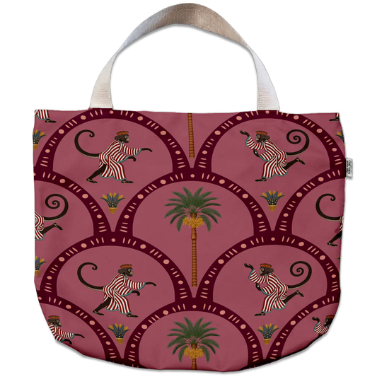A tote bag featuring a dusty rose pink background with a repeating pattern of stylized walking monkeys, palm trees, and dark red arched frames. The bag has a cream-colored fabric handle and a small side tag.