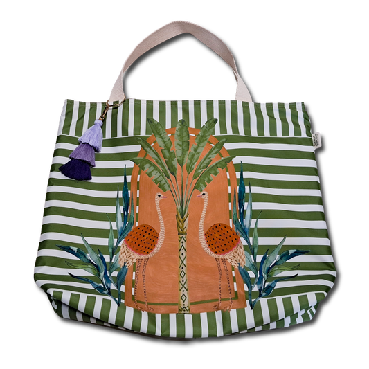 A canvas tote bag with green and white horizontal stripes, featuring a central illustration of two flamingos in front of a palm tree. The bag has light beige shoulder straps and a decorative purple tassel charm on the left side, shown against a plain white background.