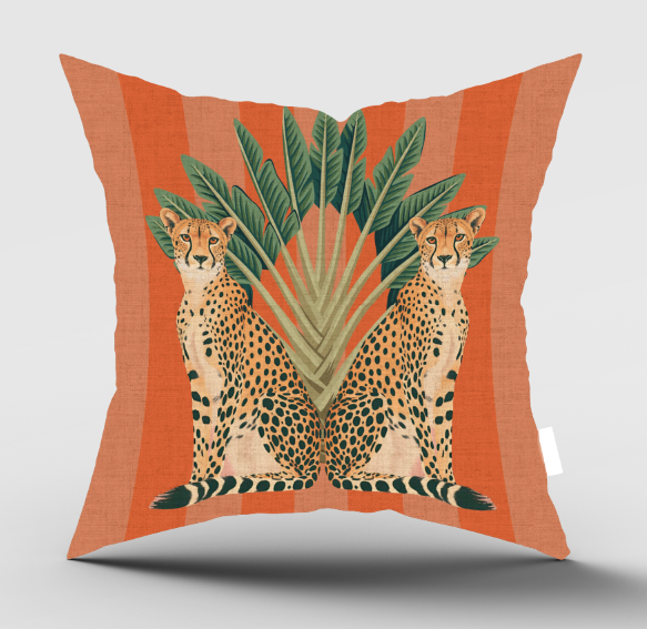 A square decorative throw pillow with a vibrant orange and peach striped background, featuring an illustration of two seated cheetahs facing each other in front of green palm leaves. A small white brand tag is visible on the right edge.