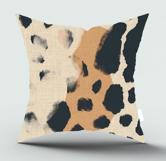 A square throw pillow with a modern abstract animal print pattern featuring black spots over a split design of beige linen and warm tan, shown on a light gray background.