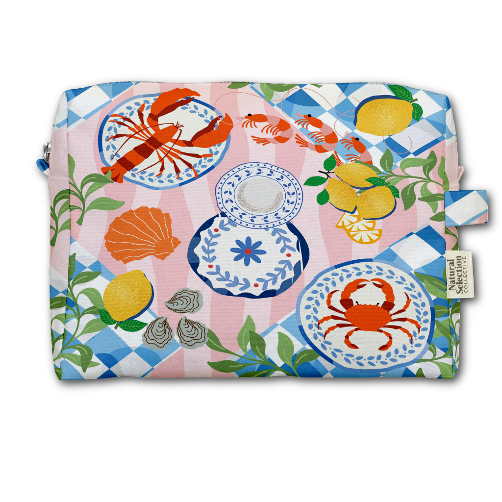 A rectangular zippered pouch with a colorful coastal print featuring a lobster, crab, lemons, seashells, fish, and blue-and-white plates on a pink and blue checkered background. A small fabric tag on the right reads Natural Living Co.