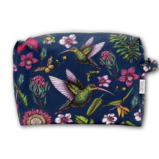 A rectangular zippered pouch with a dark navy blue background and a vibrant tropical print featuring green hummingbirds, pink flowers, and green leaves. A small white fabric tag on the right side reads 'The Art of Travel'.
