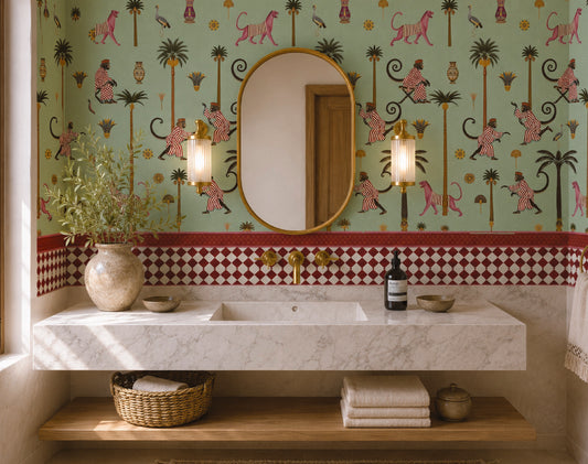A styled bathroom interior featuring a floating white marble double sink vanity with a wooden shelf below, set against a light green wallpaper with pink and black tropical animal and palm motifs, plus a red-and-white check border. An oval mirror, two wall sconces, towels, and decorative objects are visible.