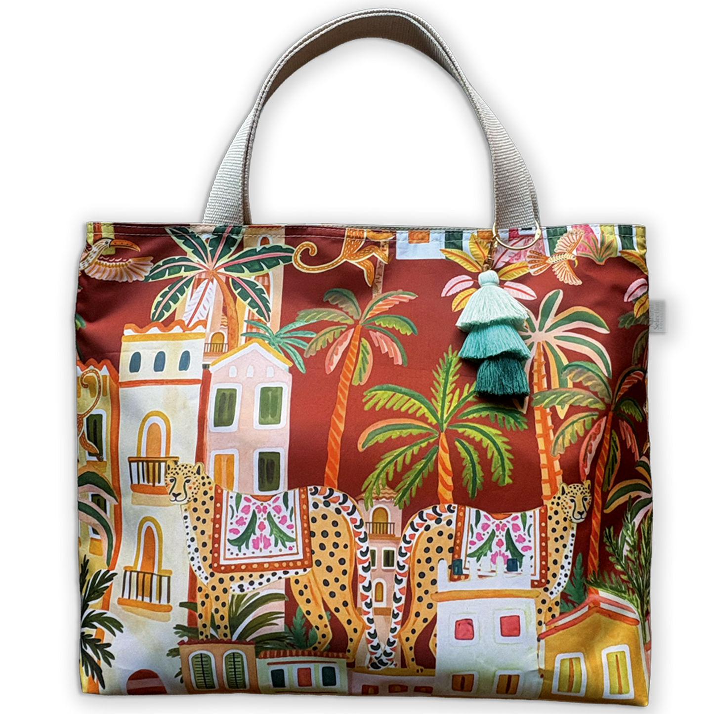 A tote bag with a vibrant, multicolor tropical and architectural print featuring palm trees, buildings, and giraffes on a dark red background. It has light gray fabric handles and a decorative tassel charm.