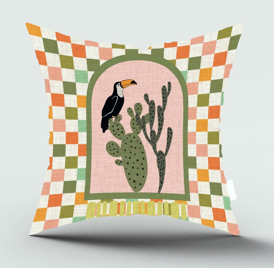 A square decorative throw pillow with a colorful geometric checkered border and a central illustration of a black-and-white toucan perched on a green cactus against a pale pink background.