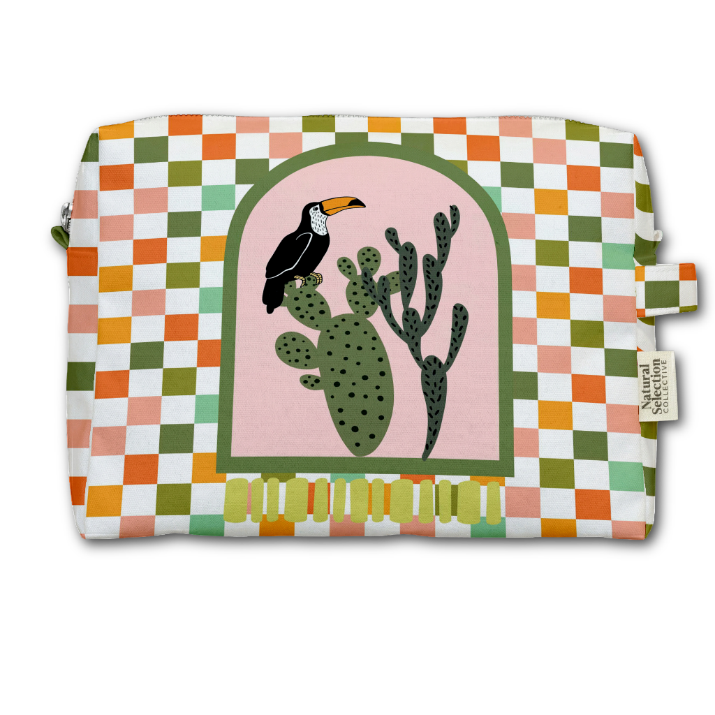 A rectangular zippered pouch with a colorful geometric checkered pattern in green, orange, pink, and white. The center features an arched illustration of a black toucan perched on a green prickly pear cactus against a light pink background.