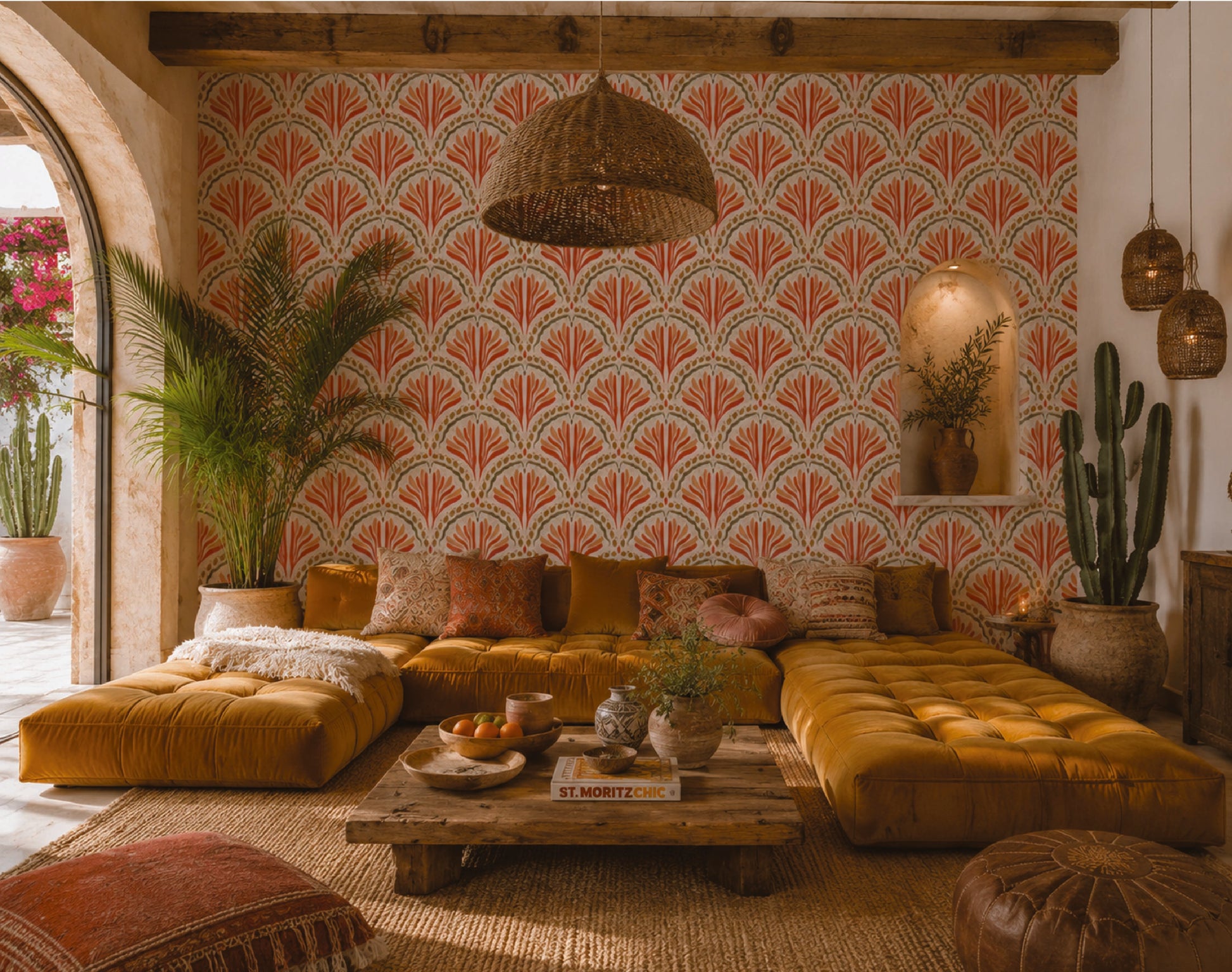 A styled bohemian living room featuring a mustard yellow tufted sectional sofa with matching pillows, a low wooden coffee table, a woven wicker pendant light, and a wall covered in red and beige fan-patterned wallpaper. The room is decorated with potted cacti, a large palm plant, and rustic wooden ceiling beams.