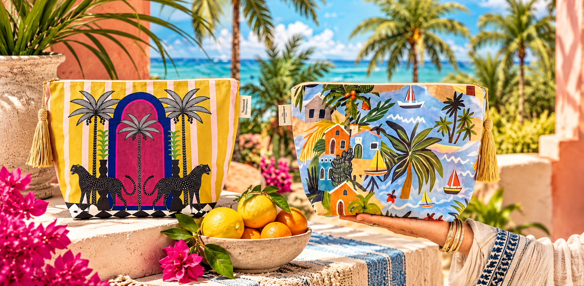 Two colorful printed pouches or bags displayed on a table outdoors, with a woman's hand holding one bag. The scene includes palm trees, ocean, flowers, lemons, and a table runner, making this a Lifestyle image.